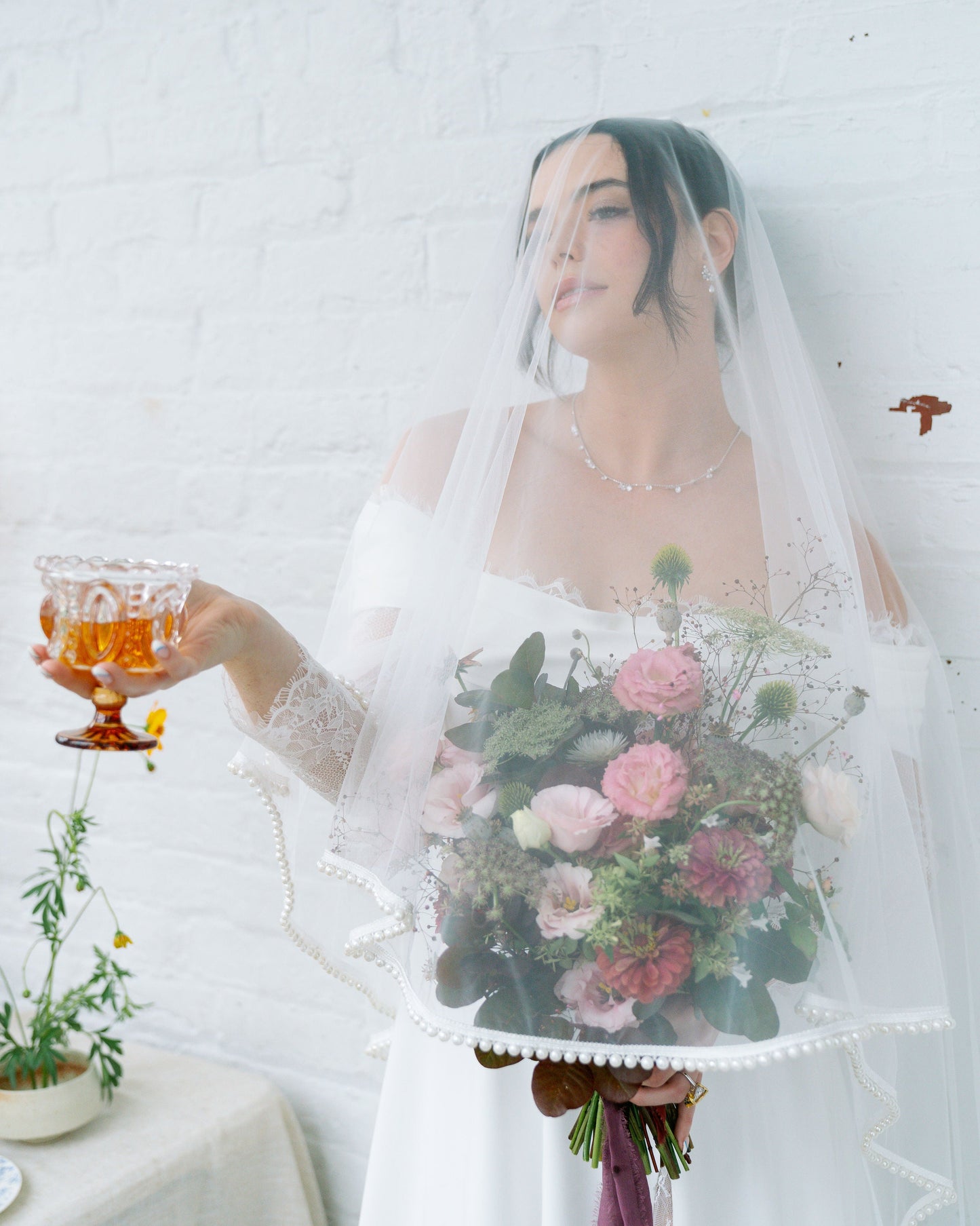 Woman in a white dress holding a bouquet of flowers and a glass with a drink, standing against a white wall.